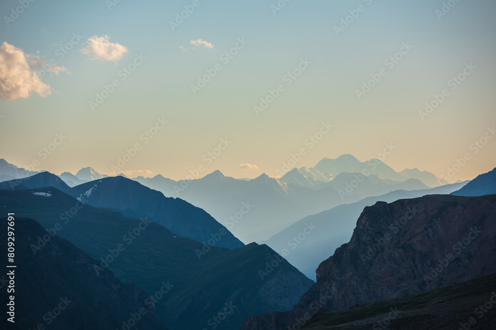 Obraz premium Deep gorge between layered silhouettes of sharp rocky ridge spurs under huge snow-capped mountain peak in golden blue gradient sky. Sheer crags of mount valley under giant ice top of gold sunset color