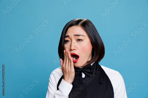 Sleepy young aisan woman receptionist yawning and covering mouth with arm. Restaurant tired overworked waitress wearing uniform feeling exhausted while posing on blue background photo