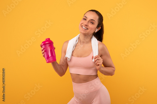 A woman wearing a pink sports bra is holding a matching pink shaker in her hand. She appears ready for a workout or fitness routine, standing against a plain background.