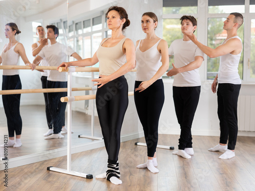 Concentrated adult male and female ballet students practicing basic positions at barre during group session under guidance of experienced instructor in well-lit choreography studio
