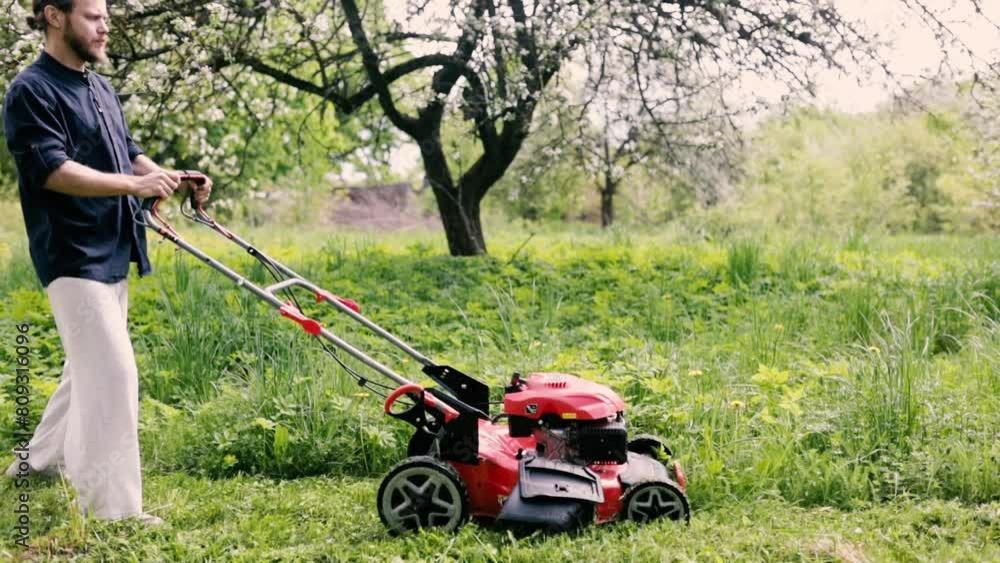 man with a lawn mower mows grass in a garden in flowering trees man with a lawn mower mows grass in a garden in flowering trees