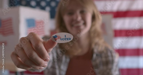 A young white woman holding up an I voted sticker at a polling station with American Flag in the background  