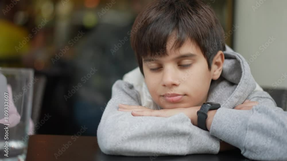 A young boy with a pensive expression rests his chin on his crossed arms atop a table, wearing a gray hoodie and gazing directly forward. A young boy with a pensive expression rests his chin on his crossed arms atop a table, wearing a gray hoodie and gazing directly forward.