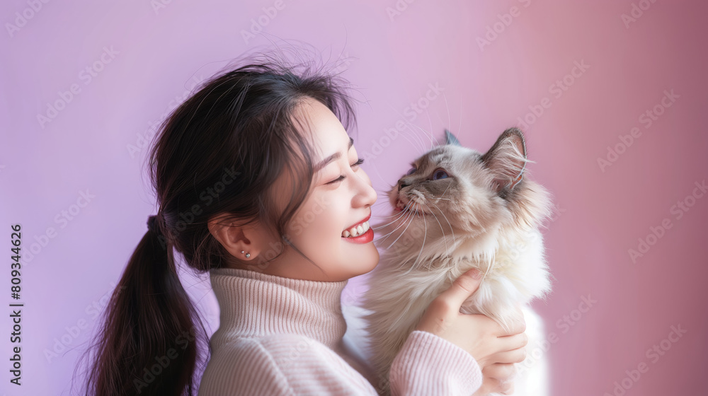 Young Asian woman enjoying a tender moment with her fluffy cat against a soft pink background. Young Asian woman enjoying a tender moment with her fluffy cat against a soft pink background.