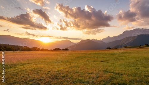 the sun is setting casting a warm afterglow over the grassy field with mountains in the background cumulus clouds dot the sky creating a picturesque natural landscape
