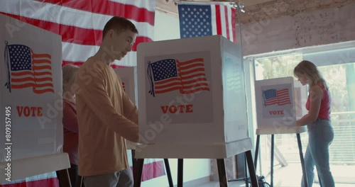 Low angle of a group of diverse and focused voters voting at a polling station with sliding glass doors, American flags and trees in the background
