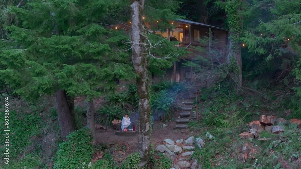 A girl unwinds by a crackling fire outside a cozy wooden cabin nestled in a lush forest near a river at dusk. Washington. USA. Aerial. 4K footage. A girl unwinds by a crackling fire outside a cozy wooden cabin nestled in a lush forest near a river at dusk. Washington. USA. Aerial. 4K footage.
