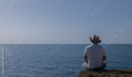 Rear view of adult man in hat sitting on hill against sea and sky