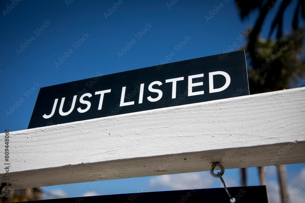 A black realtor sign with white letters advertising a new home listing  Plam trees are in the background
