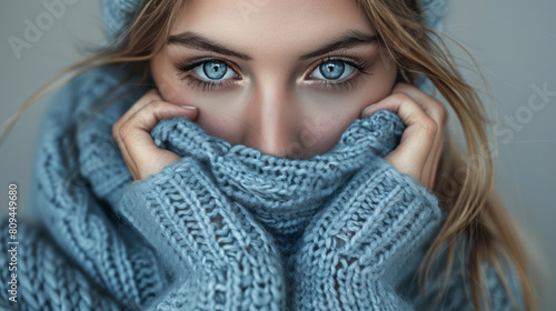 A young woman in a blue sweater covers her face with her hands, feeling cold and hugging herself against a grey background, looking at the camera while wearing a big knitted pullover. photo