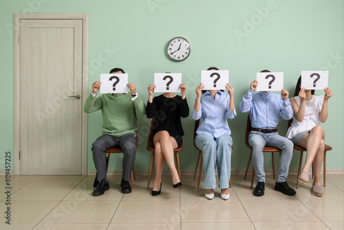 Young people holding paper sheets with question marks while waiting for job interview in room