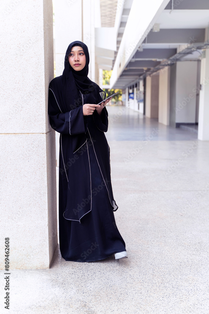 Young Asian beautiful woman Muslim student portrait in traditional clothes relaxing and hold tablet on school building background. Young Asian beautiful woman Muslim student portrait in traditional clothes relaxing and hold tablet on school building background.