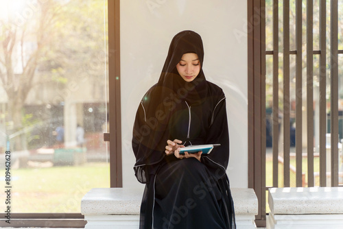 Canvas Print Asian Muslim wearing traditional clothes sit and review the lesson on tablet for the exam in university with sun fare and blurred background