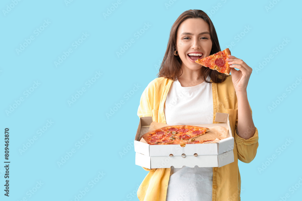 Young woman eating piece of tasty pizza and holding cardboard box on blue background Young woman eating piece of tasty pizza and holding cardboard box on blue background
