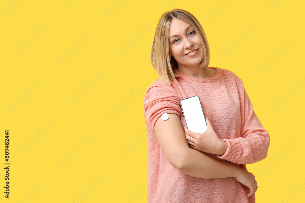 Woman with glucose sensor for measuring blood sugar level and phone on yellow background. Diabetes concept Woman with glucose sensor for measuring blood sugar level and phone on yellow background. Diabetes concept