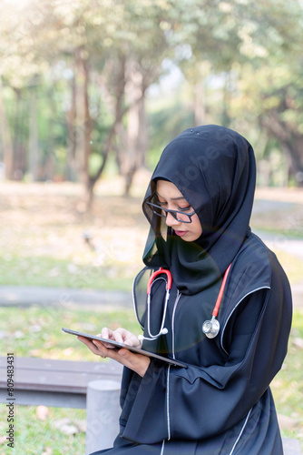 Young Asian beautiful woman Muslim medical student wearing traditional clothes relaxing with tablet in the hospital park with sun fare and blurred background. © Atiwat