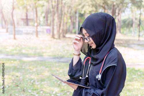 Closeup young Asian beautiful woman Muslim doctor wearing traditional clothes relaxing with tablet in the hospital park with sun fare and blurred background. © Atiwat