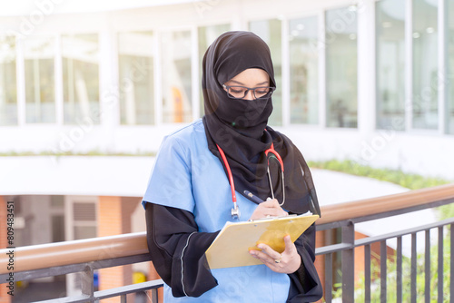 Female Muslim doctor in Abaya niqab traditional clothes works at modern clinic office checking of work list on clipboard with sun fare background. © Atiwat