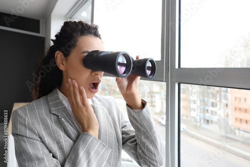 Canvas Print Shocked young African-American businesswoman looking through binoculars in windo