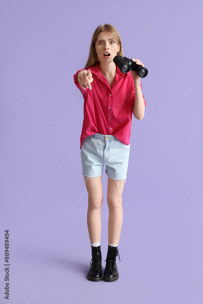 Shocked young woman with binoculars pointing at viewer on lilac background Shocked young woman with binoculars pointing at viewer on lilac background