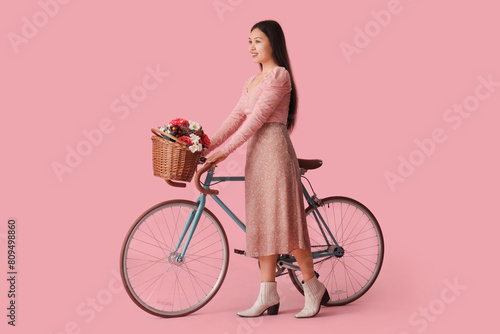 Beautiful young Asian woman with bicycle and bouquet of flowers on pink background © Pixel-Shot