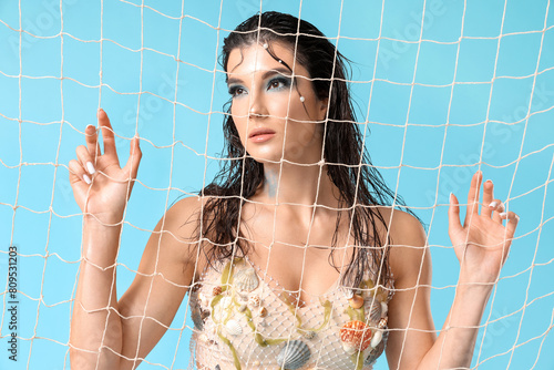 Beautiful young woman dressed as mythical mermaid visible through net on blue background photo