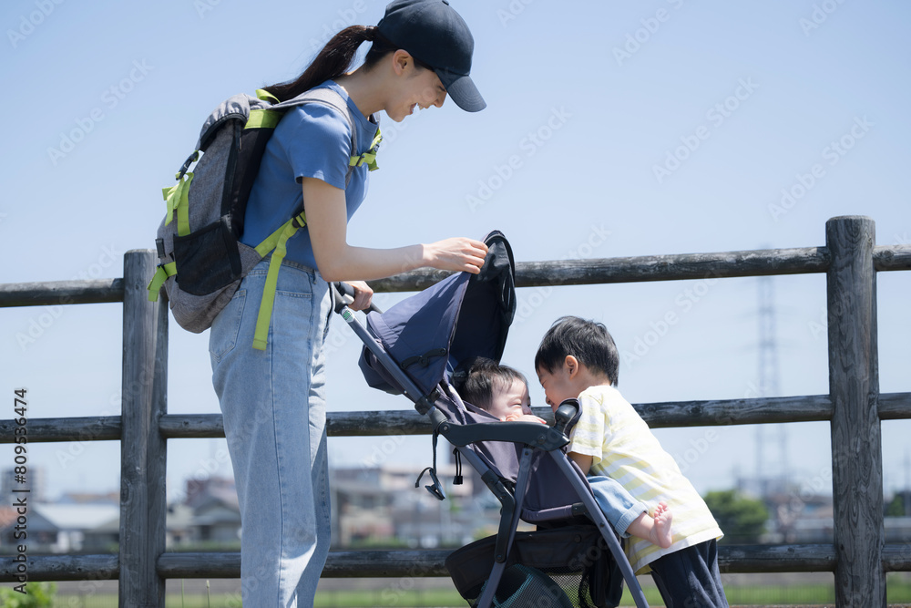Obraz premium 川沿いの河川敷でベビーカーを押す母親と下の子の面倒やお世話をするかわいい兄