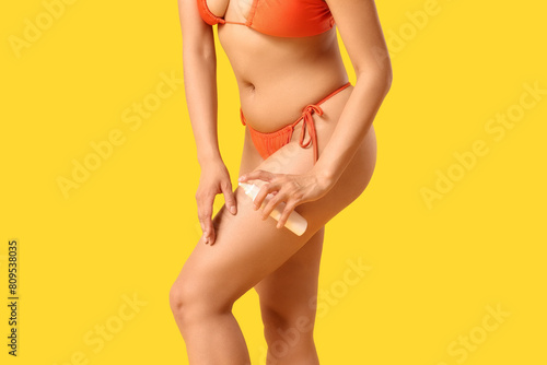 Young African-American woman in swimsuit applying sunscreen cream on her leg against yellow background