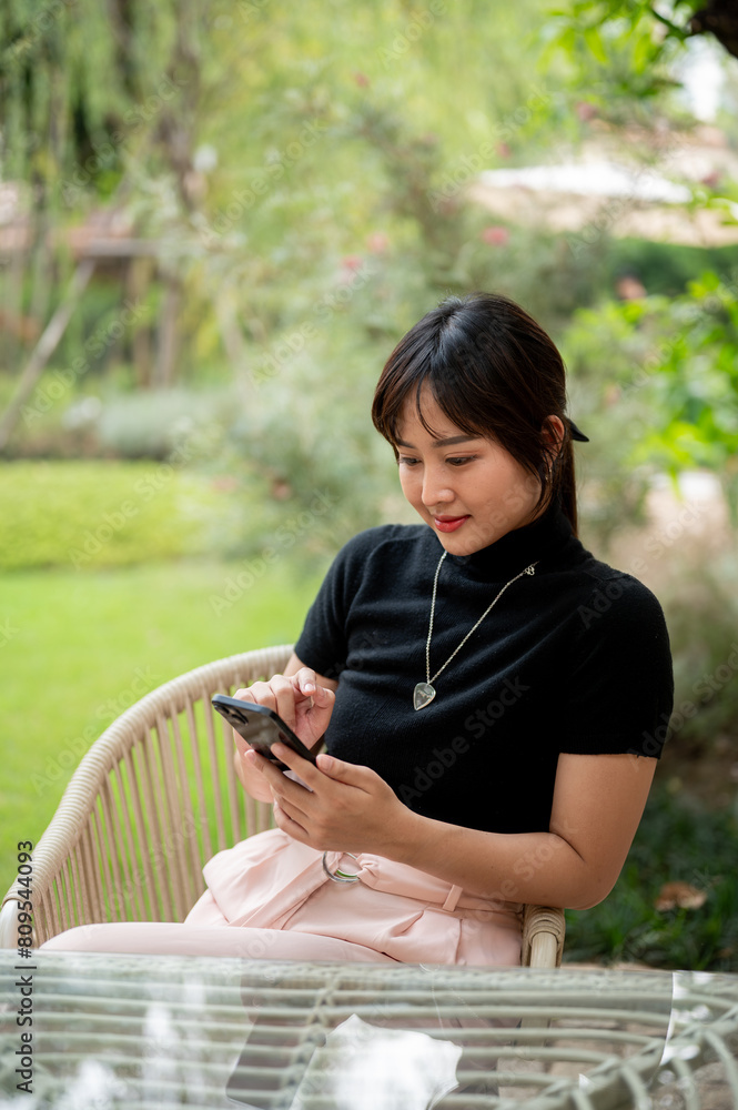A beautiful Asian woman sits at a table in a green garden using her smartphone, checking messages. A beautiful Asian woman sits at a table in a green garden using her smartphone, checking messages.