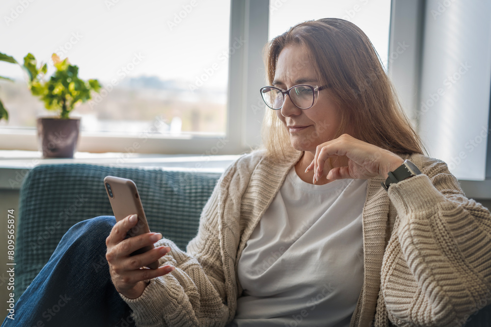 Caucasian attractive mature middle aged woman sitting on the couch at home using smartphone