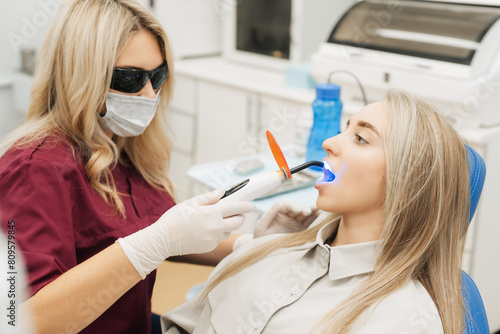 Professional female doctor uses ultraviolet lamp after making a filling on a tooth for joyful happy caucasian woman in modern dental cabinet