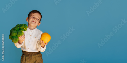 A studio shot of a boy holding fresh broccoli and an orange on a blue background with a copy of the space. The concept of healthy baby food. photo