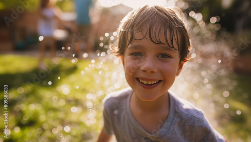 Lachender Junge im Garten spielt mit Wasserspritzern, Familie im unscharfen Hintergrund, Abkühlung und Spaß im Sommer 