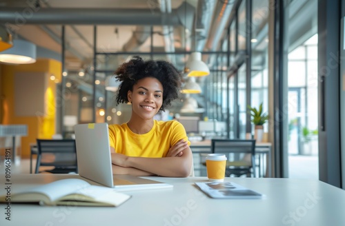 Free Download A woman in a yellow shirt is sitting at a desk with a laptop and a cup of coffee. She is smiling and she is enjoying her time at work,Sunny Workdays: A Woman's Joyful Moment at the Desk - GFXLoad