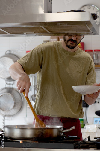 man slightly obscured, almost unrecognizable by the stainless steel fume hood, stirring a pan, green caci t-shirt, pots hanging in the background, kitchen environment. photo