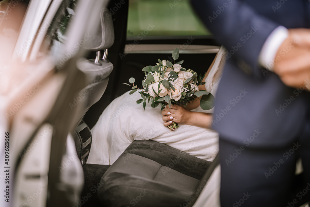 Valmiera, Latvia - August 25, 2023 - a bride in a white dress holding a bouquet of flowers inside a car, with a man in a suit visible nearby. Valmiera, Latvia - August 25, 2023 - a bride in a white dress holding a bouquet of flowers inside a car, with a man in a suit visible nearby.