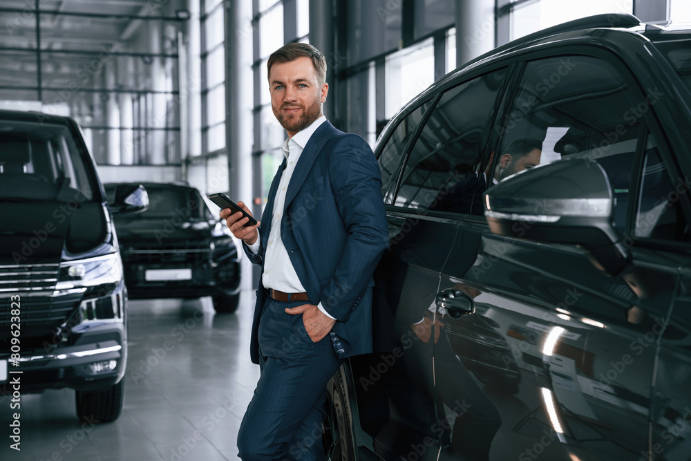 Leaning on the vehicle and holding smartphone. A businessman is in a car dealership Leaning on the vehicle and holding smartphone. A businessman is in a car dealership