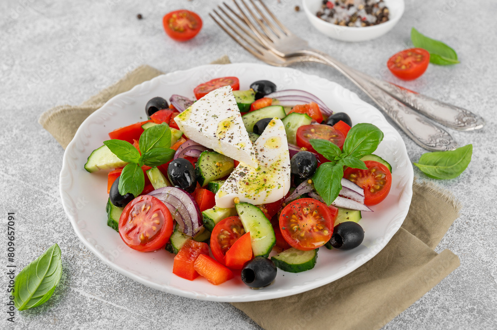 Greek salad of fresh juicy vegetables, feta cheese, herbs and olives on a white plate on a light concrete background. Healthy food. Copy space, top view.