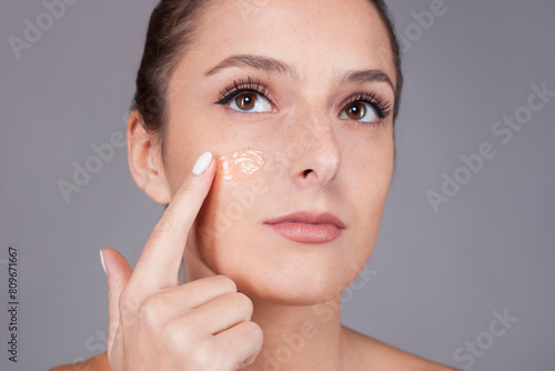 Young beautiful woman with healthy skin taking care of face with foundation isolated on gray background looking away. photo