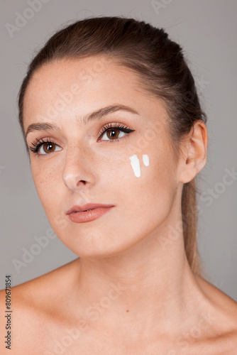Young beautiful woman with healthy skin taking care of face with cream isolated on gray background looking away. Moisturize, cosmetic, skincare concept. photo