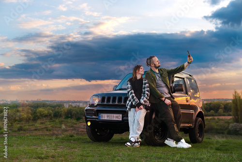 Travelers Couple. Road Trip, Man and Woman on Journey Near Their SUV off road Car Over Beautiful Landscape. Sunset clouds. taking selfie photos on a smartphone