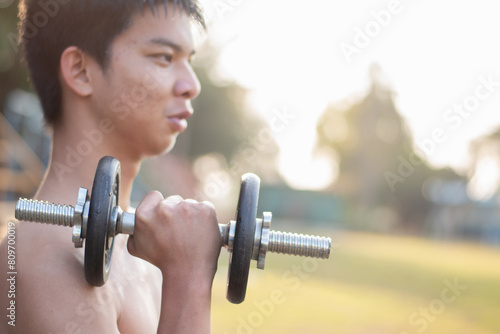 Asian preteen boy lifting dumbbells to loose weight and to do the bodybuild in grass field in late afternoon of the day, soft focus.	 photo