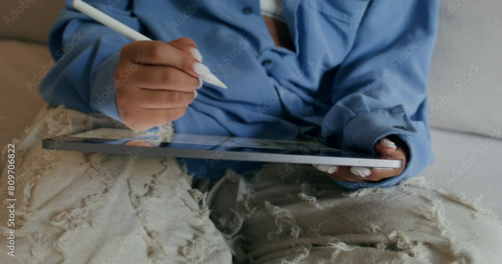 A woman uses a tablet sitting on the couch at home. She draws on it with a stylus. Close-up of her hands, an unrecognizable person A woman uses a tablet sitting on the couch at home. She draws on it with a stylus. Close-up of her hands, an unrecognizable person