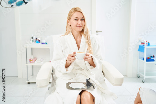 Beautiful woman in white bathrobe drinking tea in aesthetic medicine clinic
