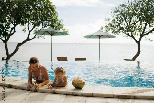 Portrait of young mother sharing coconut water with her little daughter while sitting near swimming pool with ocean view on summer vacation