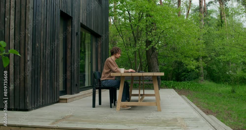 A man in casual clothes uses a laptop and drinks coffee sitting at a table on the terrace of a stylish cottage A man in casual clothes uses a laptop and drinks coffee sitting at a table on the terrace of a stylish cottage