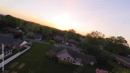 Smooth drone flight over villas and houses in suburb neighborhood in United States during golden sunset. Beautiful idyllic landscape with trees in spring season. Roofing over real estate building.