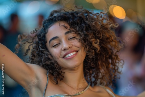 A close-up of a young woman with curly hair smiling joyfully in front of a blurred audience while dancing