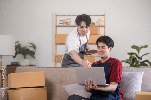 A gay couple running a small business takes order on a laptop and sells product online together in a room full of boxes.