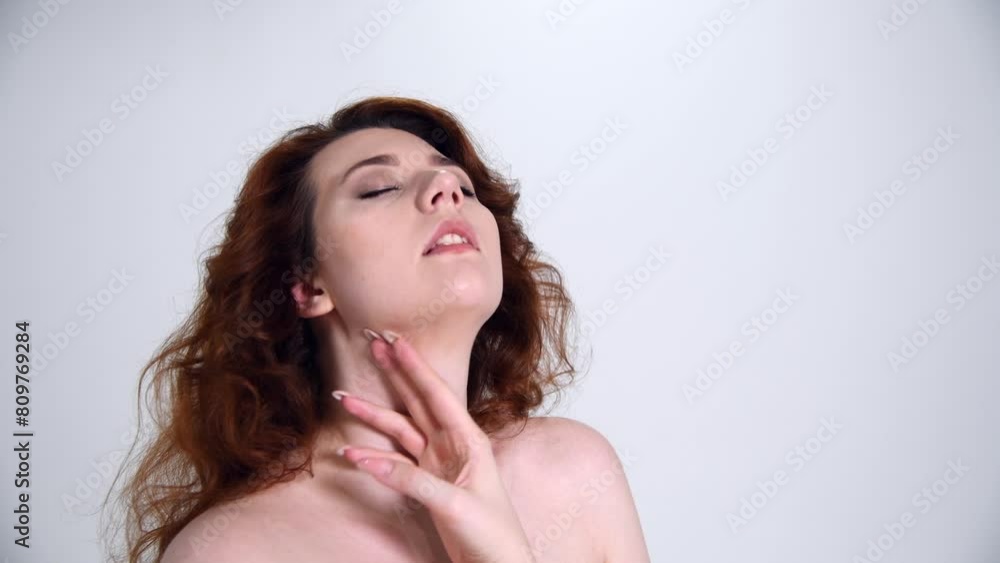 Close-up portrait of beautiful caucasian plus size woman with long curly brown hair and naked shoulders touching her skin against grey background. Real time handheld video. Beauty and skincare theme. Close-up portrait of beautiful caucasian plus size woman with long curly brown hair and naked shoulders touching her skin against grey background. Real time handheld video. Beauty and skincare theme.